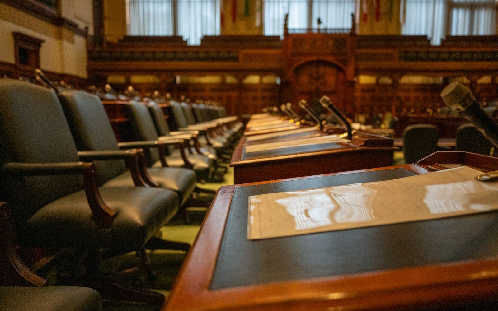 Empty leather chairs and wooden desks with microphones line a legislative chamber