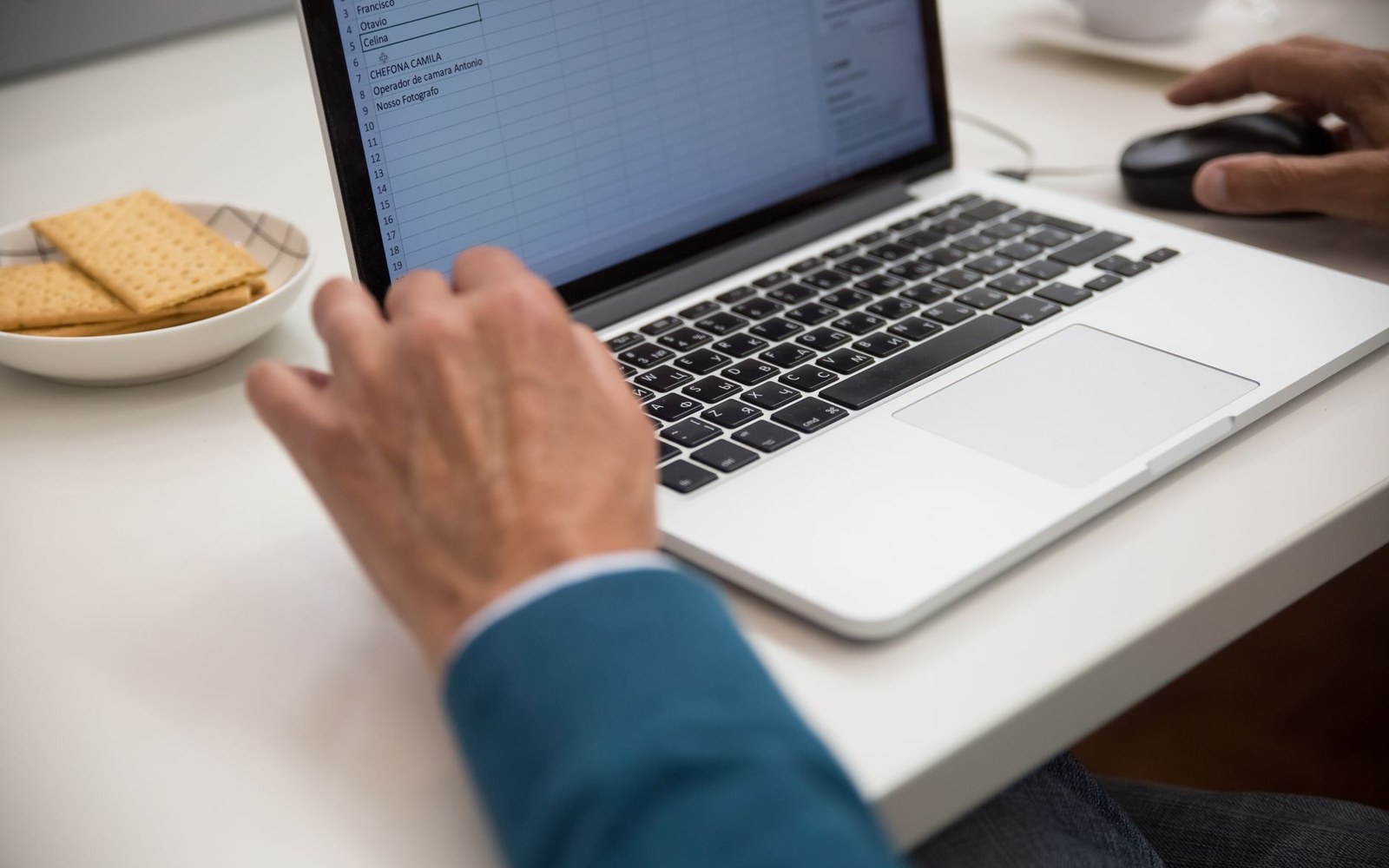 An adult's hands at a keyboard, a spreadsheet of monthly income figures on screen, coffee cup at edge of frame